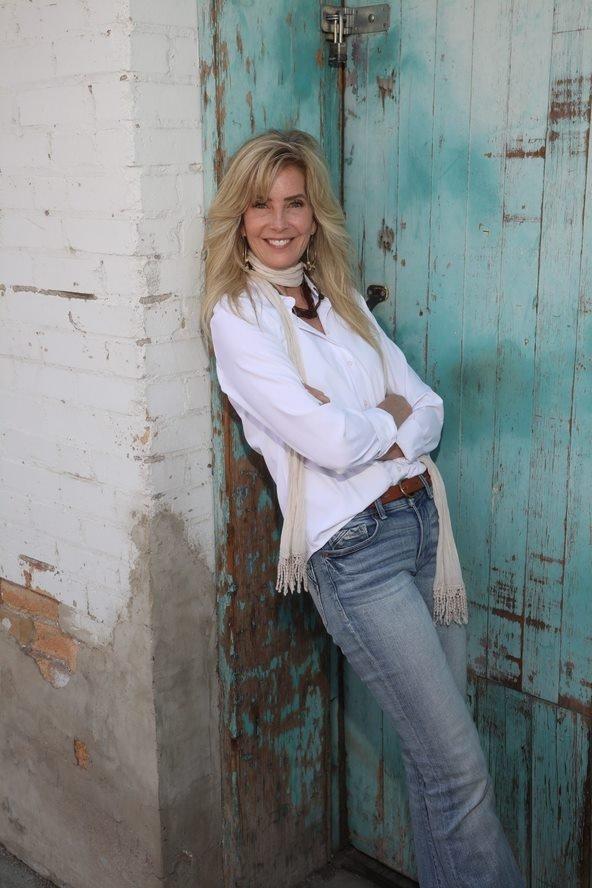 Julie Write-Leggett, a blonde woman in a white dress shirt and jeans smiling with her arms crossed and leans against a barn door that is painted sky blue
