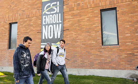 Three students walking on campus past an enroll now banner