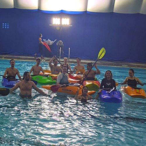 A group of people in a pool, some of them are in kayaks while others are wading.
