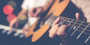 A close up image of a person playing a guitar with another person playing the piano in the background